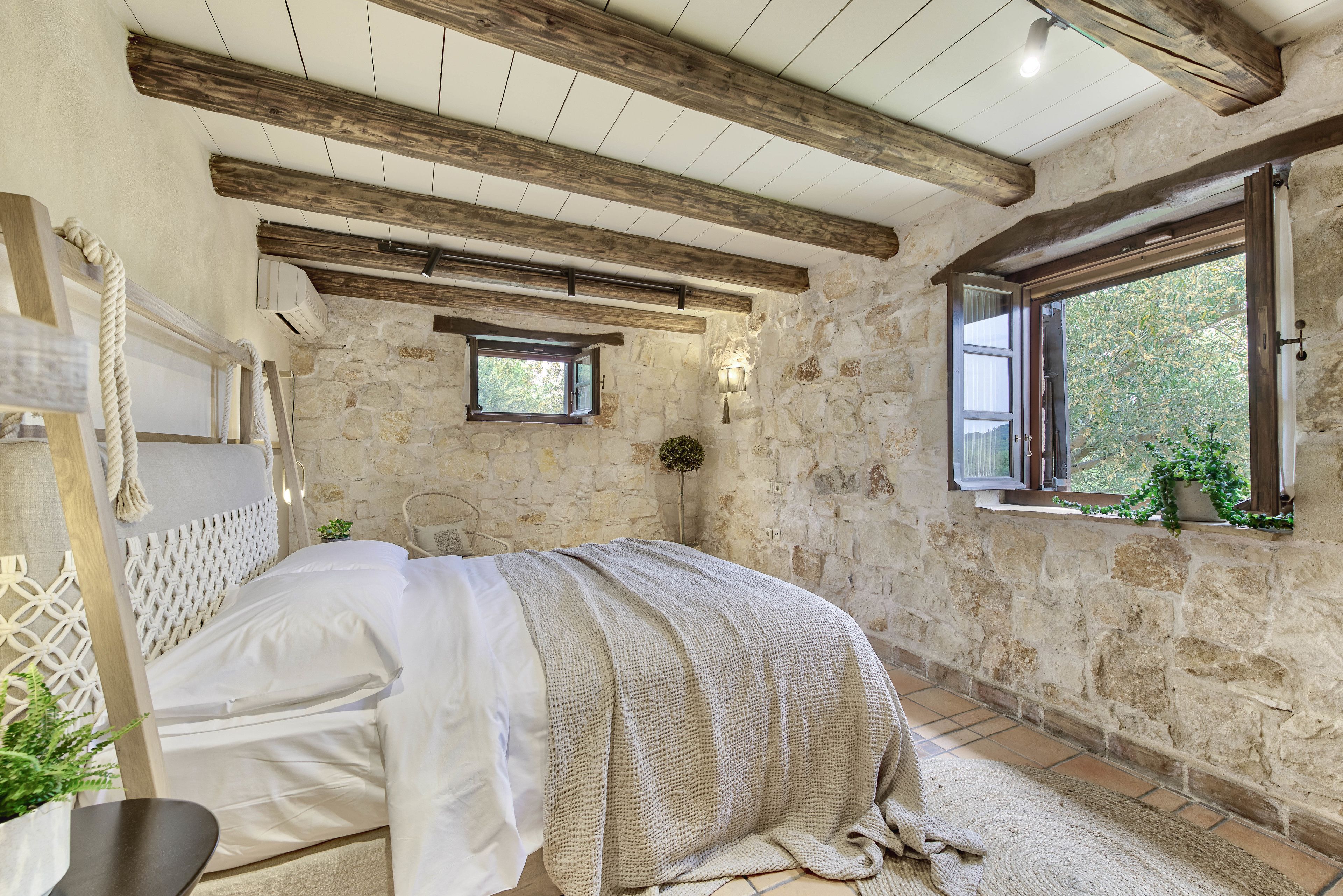 Un dormitorio con paredes de piedra, techo de madera, cama con colcha blanca, una ventana pequeña con vistas y una planta en maceta.
