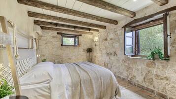 Un dormitorio con paredes de piedra, techo de madera, cama con colcha blanca, una ventana pequeña con vistas y una planta en maceta.