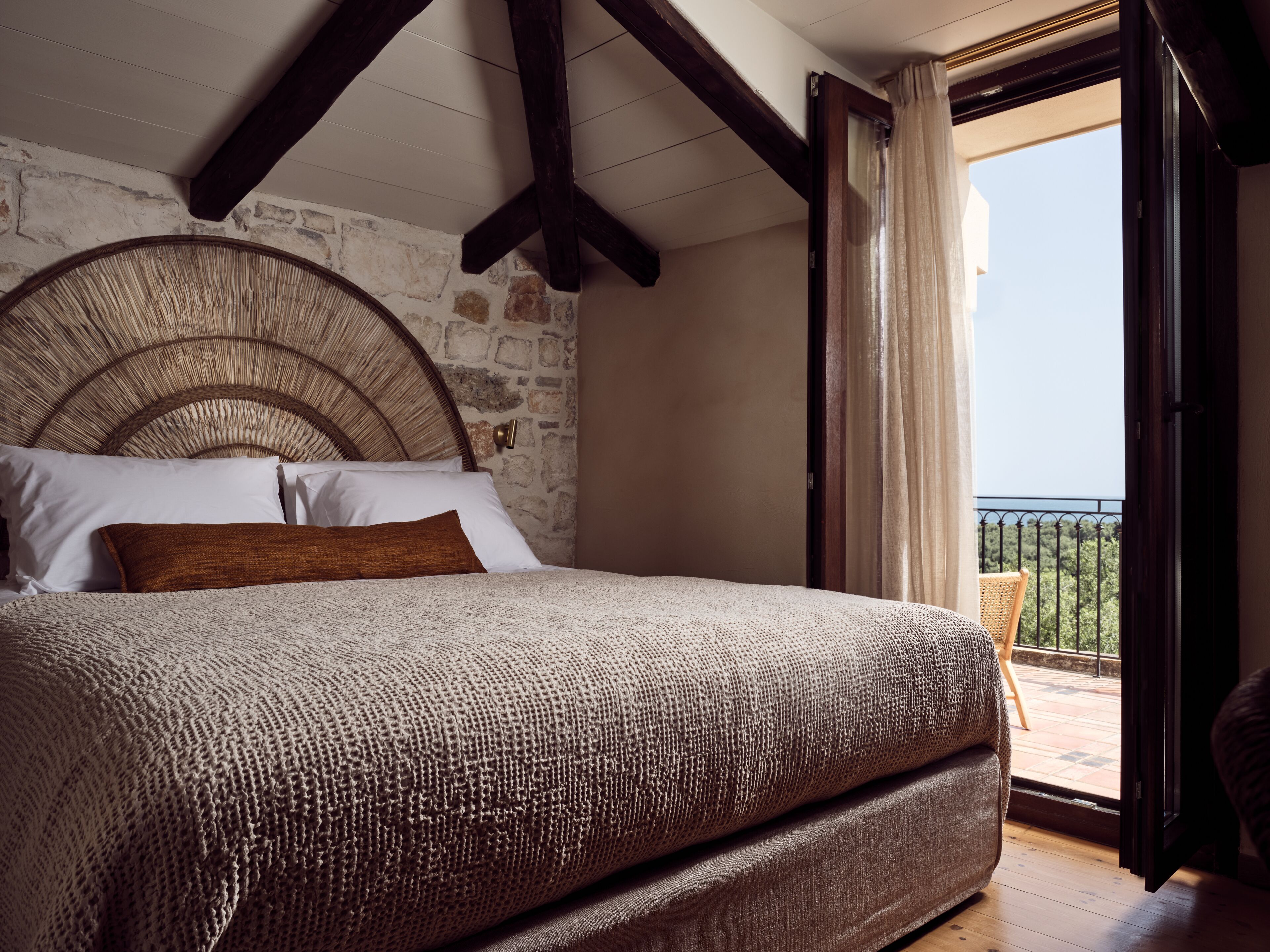 A bedroom with a stone wall, a large bed with a woven headboard, and a balcony with a view.