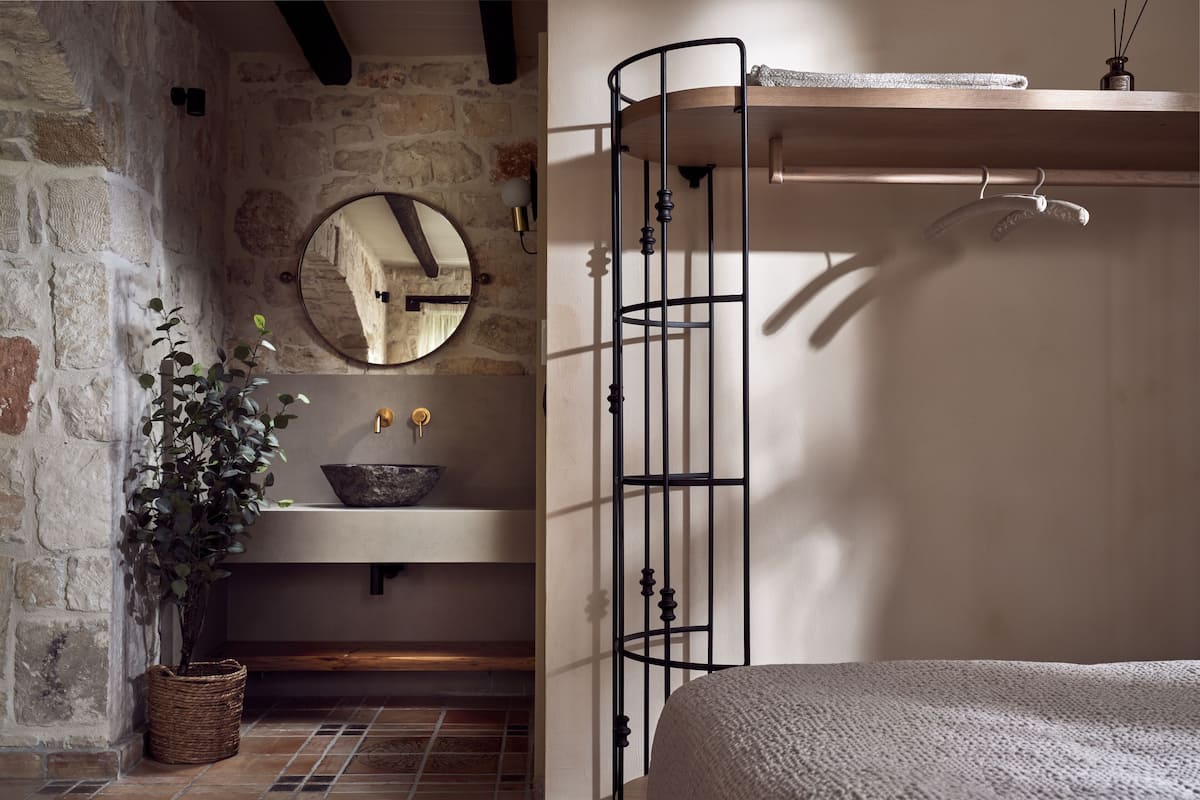 A rustic bathroom with a stone wall, a round mirror, a wooden shelf, and a metal rack.