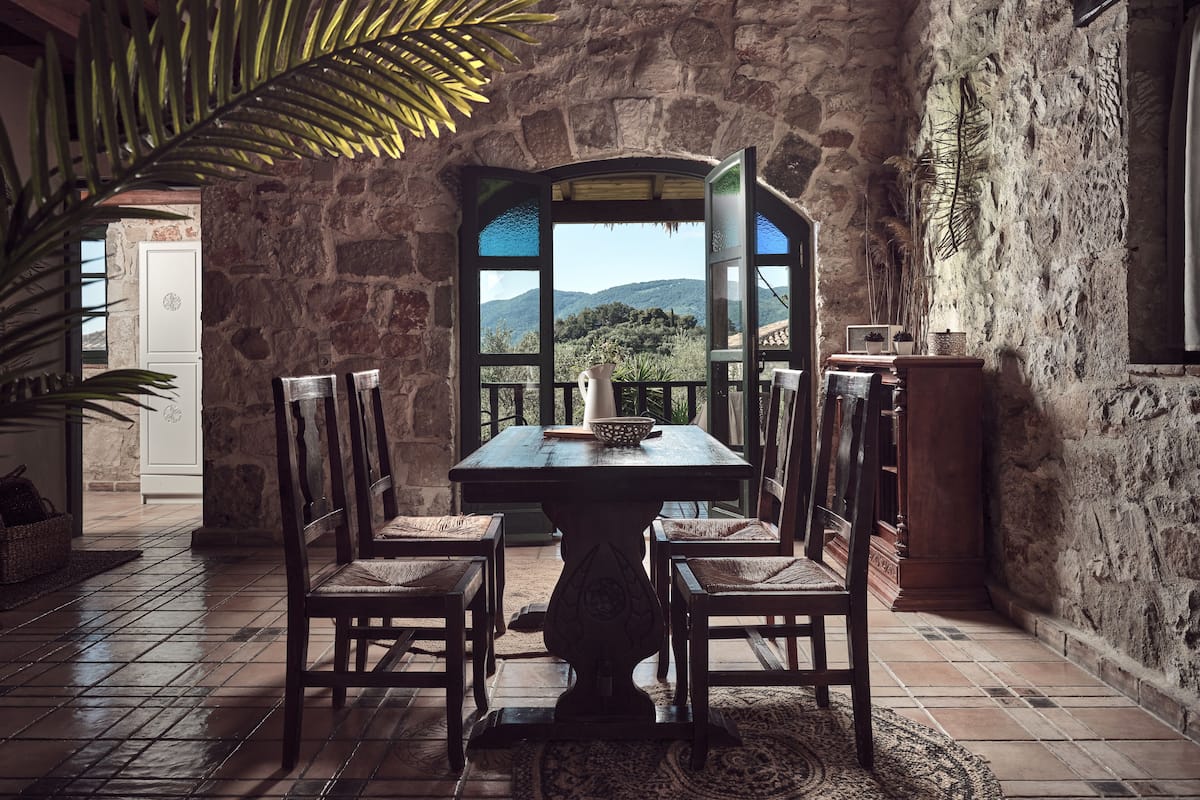 A rustic stone interior with a wooden table and chairs, a view of mountains through a window, and a potted plant.