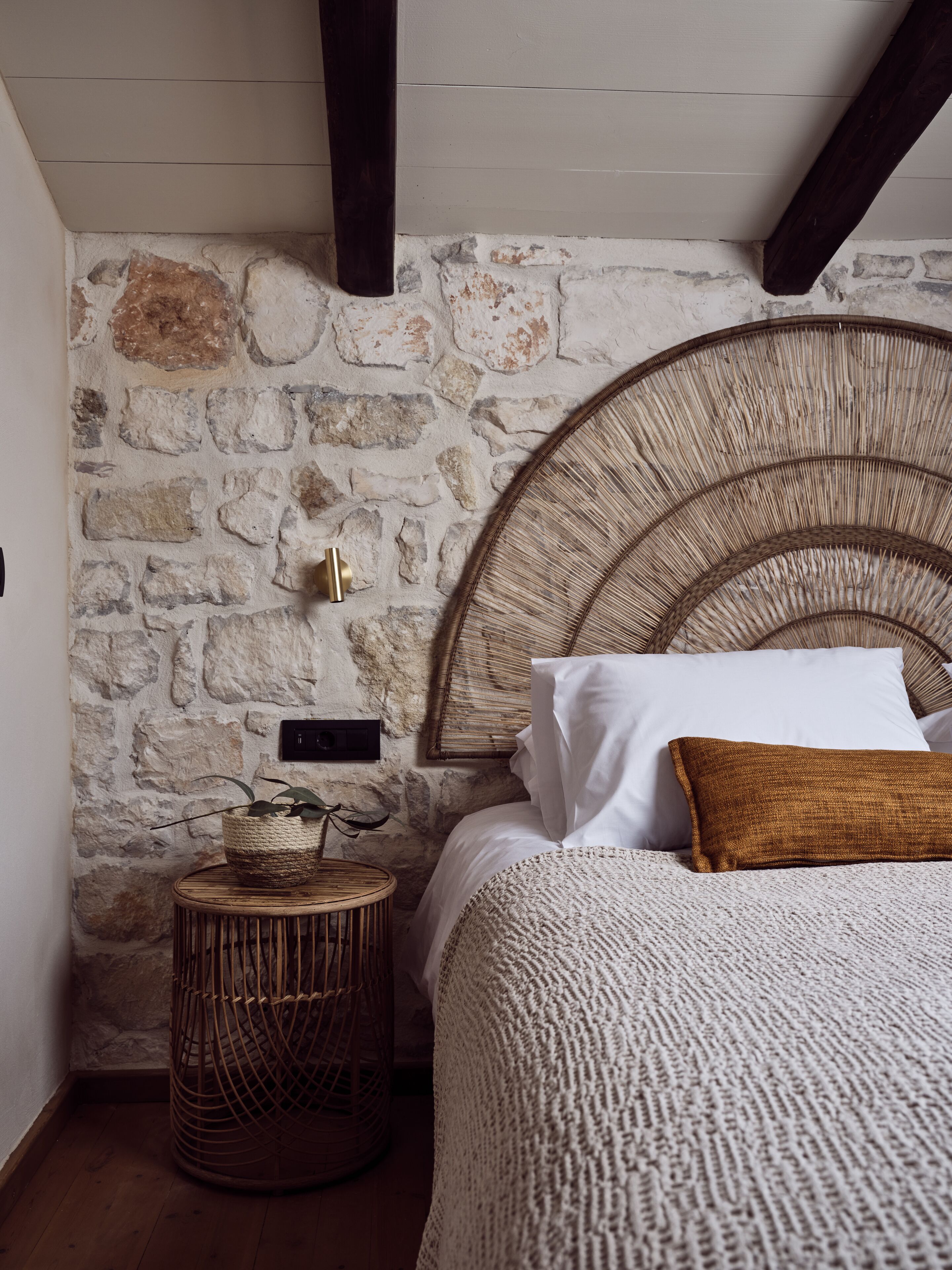 Een kamer met een stenen muur, een geweven hoofdeinde, een rond houten bijzettafeltje en een bed met wit en bruin beddengoed.