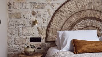 Een kamer met een stenen muur, een geweven hoofdeinde, een rond houten bijzettafeltje en een bed met wit en bruin beddengoed.