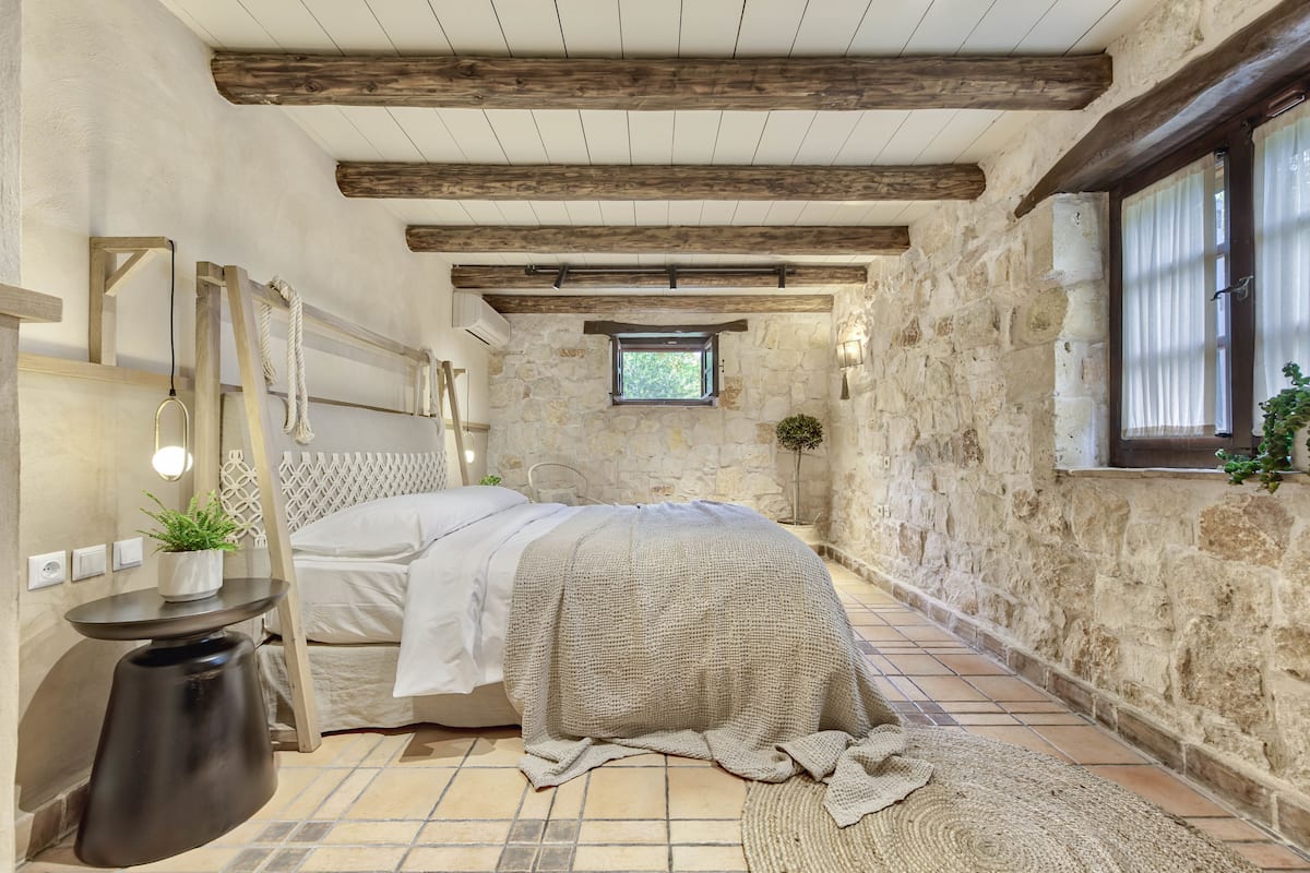 A bedroom with a stone wall, wooden beams, a bed with a white bedspread, a small table with a plant, and a window with curtains.