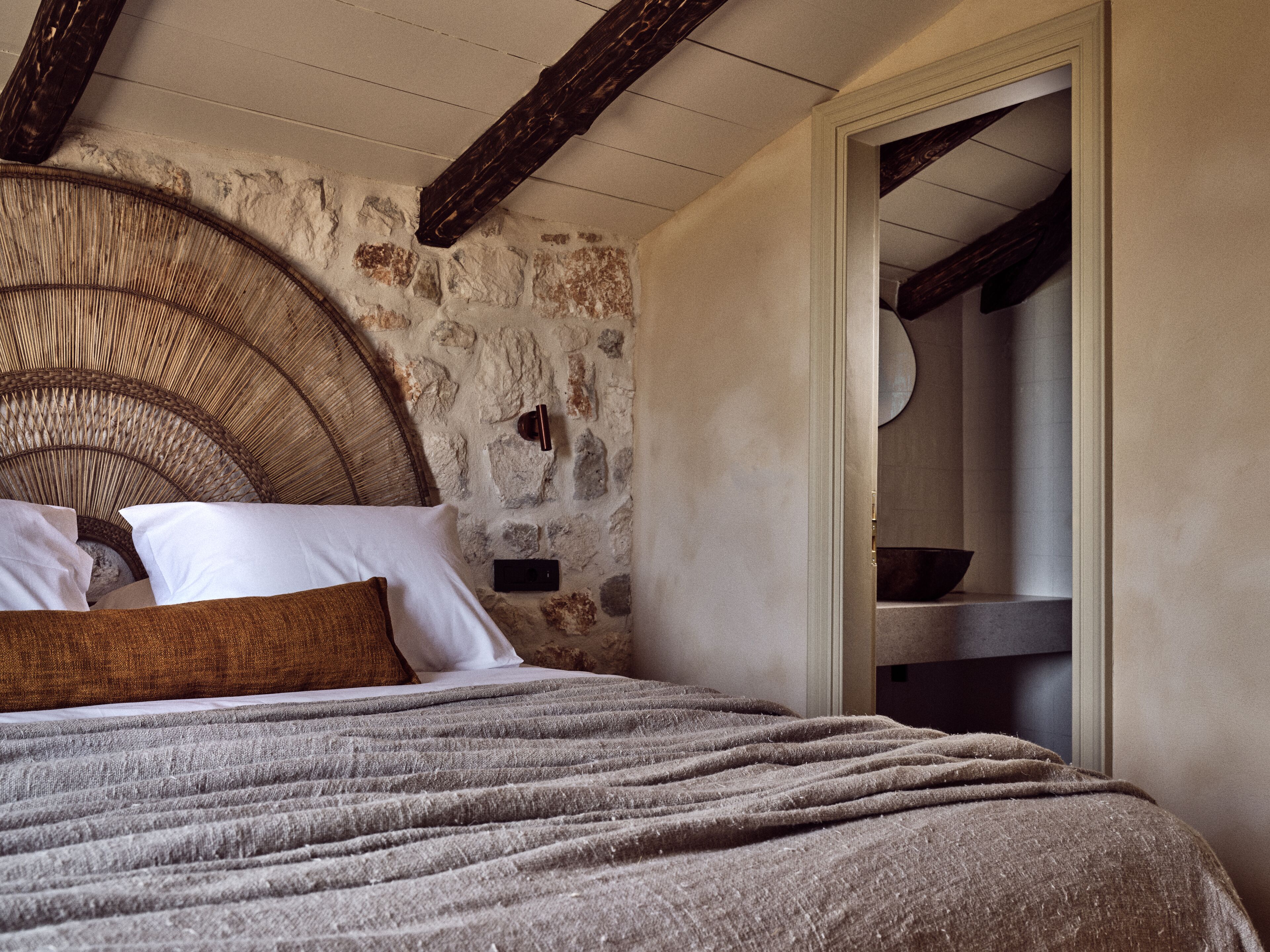 A bedroom with a stone wall, a wicker headboard, a bed with a grey blanket, and a wooden door.
