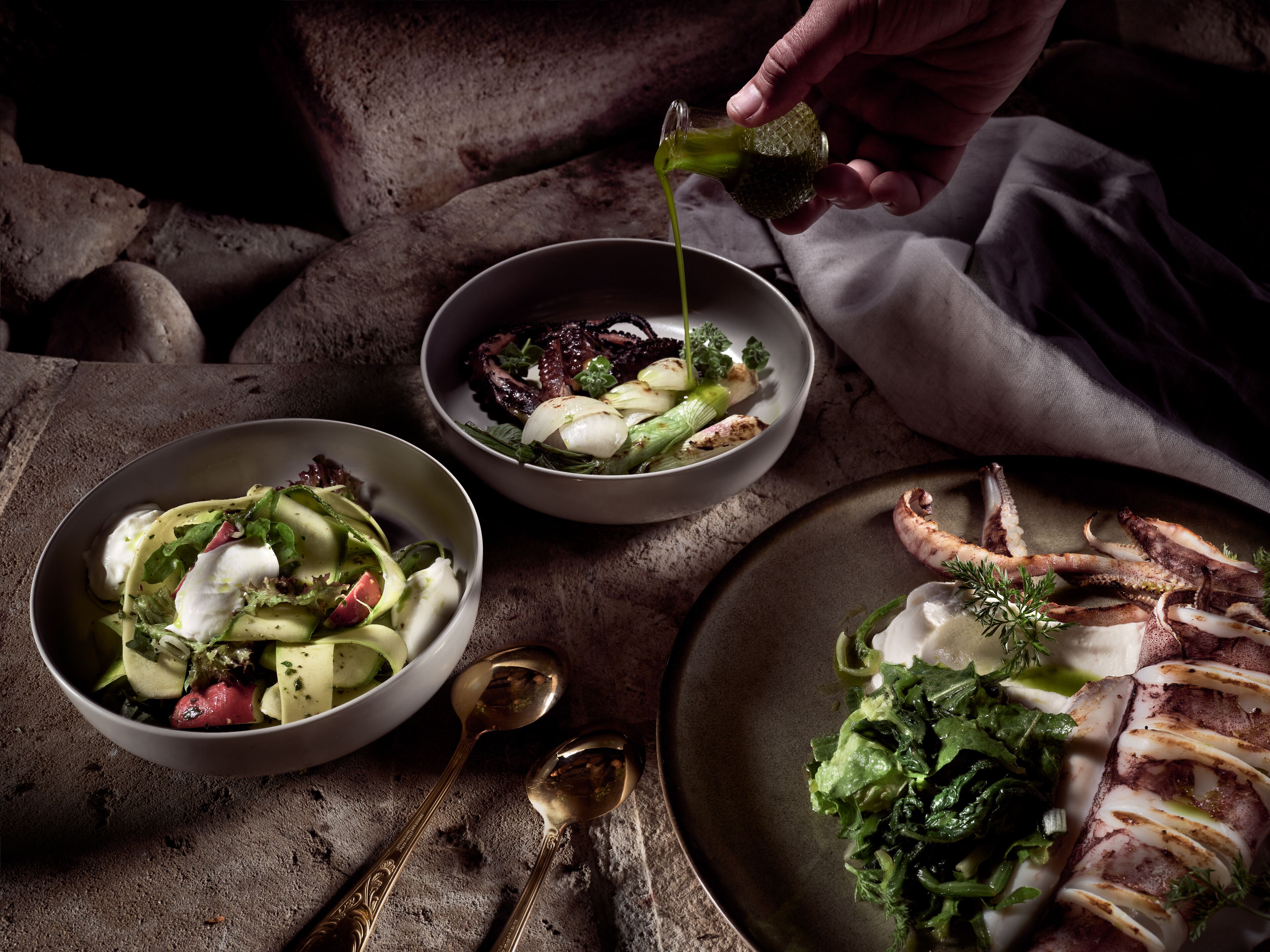 Three bowls of food on a table with spoons and a hand pouring dressing.