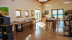 Amplia zona de comedor con techos altos y ventanas grandes, que presenta un sistema de buffet con diversas opciones de comida y diferentes disposiciones de mesas.