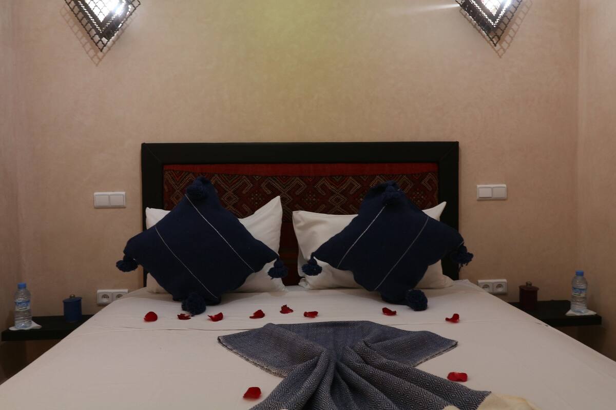A neatly made bed with a patterned blanket, two pillows, and scattered rose petals.