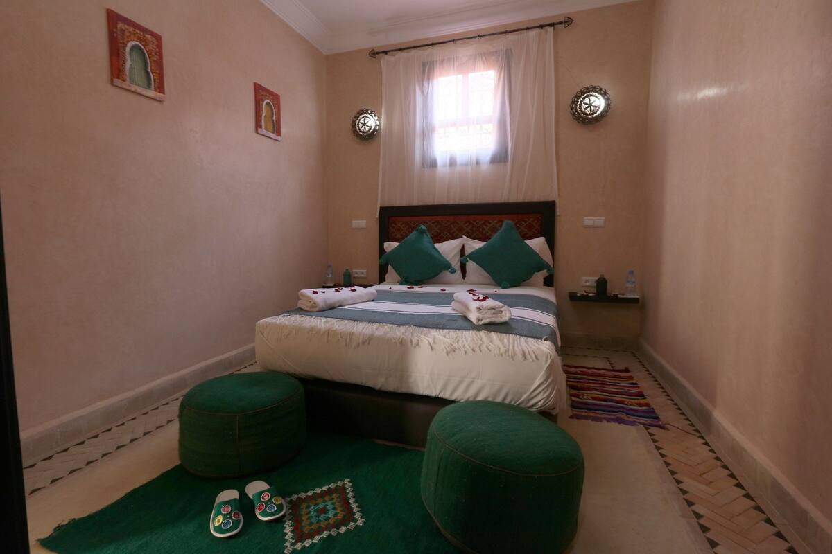 A small, cozy bedroom with a bed, two green ottomans, a patterned rug, and a small side table with a water bottle.