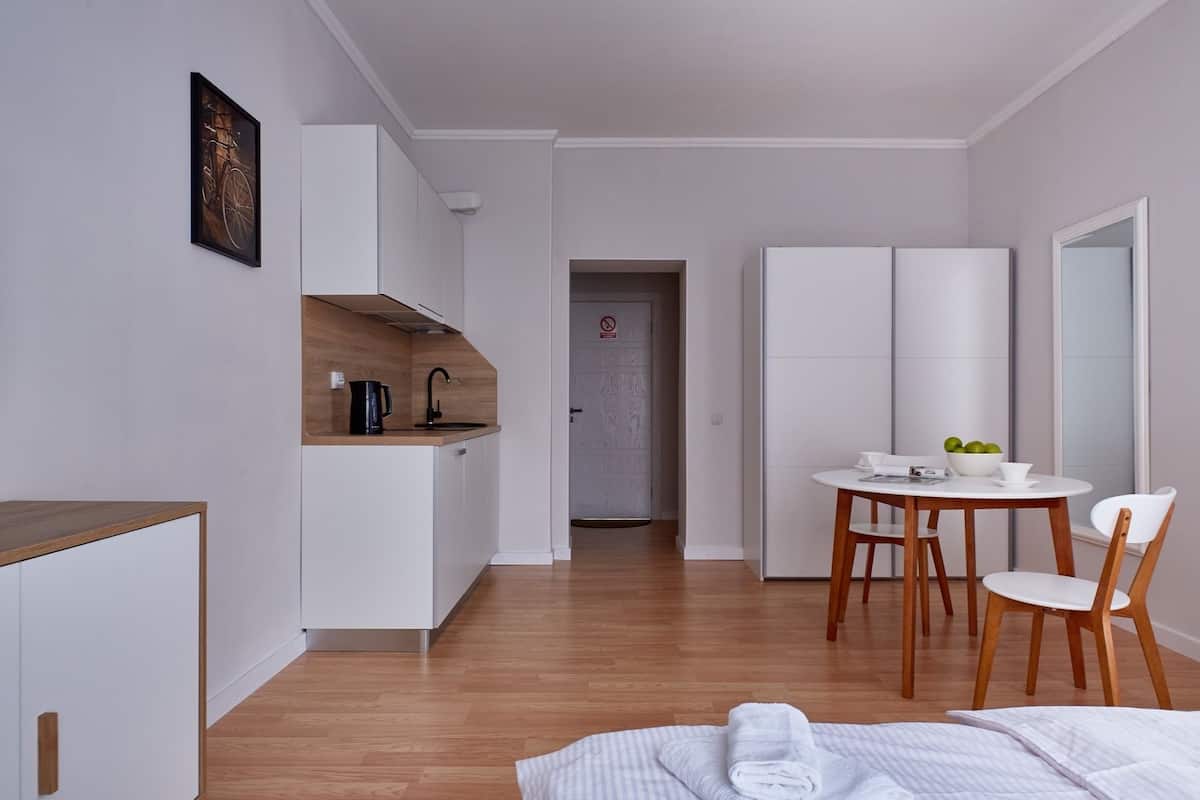 A modern, minimalist hotel room with a wooden floor, a bed with white linens, a small kitchen area with a coffee maker, and a dining table with chairs.