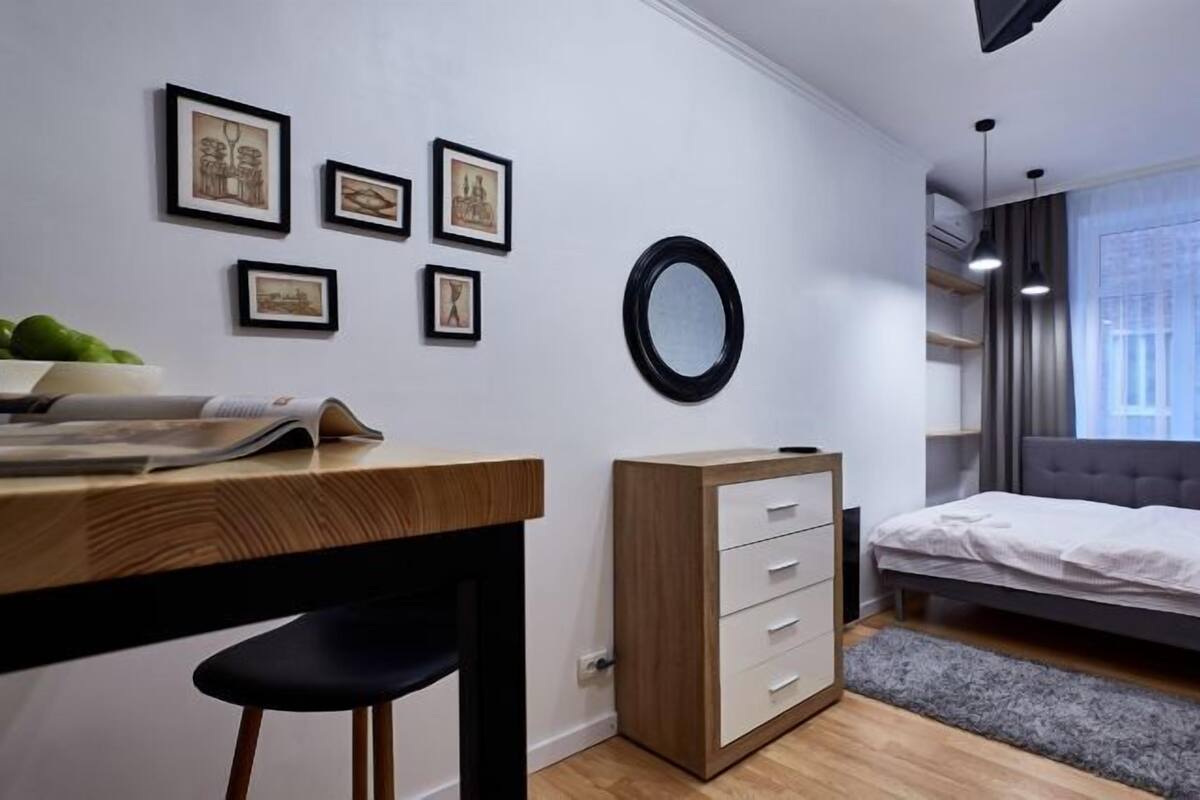 A modern bedroom with a bed, a desk, and a chair. There are framed pictures on the wall and a round mirror.