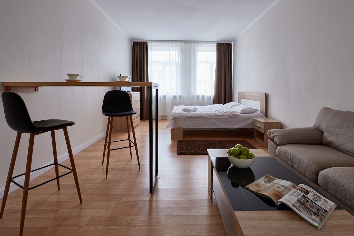 A modern hotel room with a wooden desk, two black chairs, a bed with white linens, a brown sofa, and a coffee table with a bowl of fruit and a magazine.
