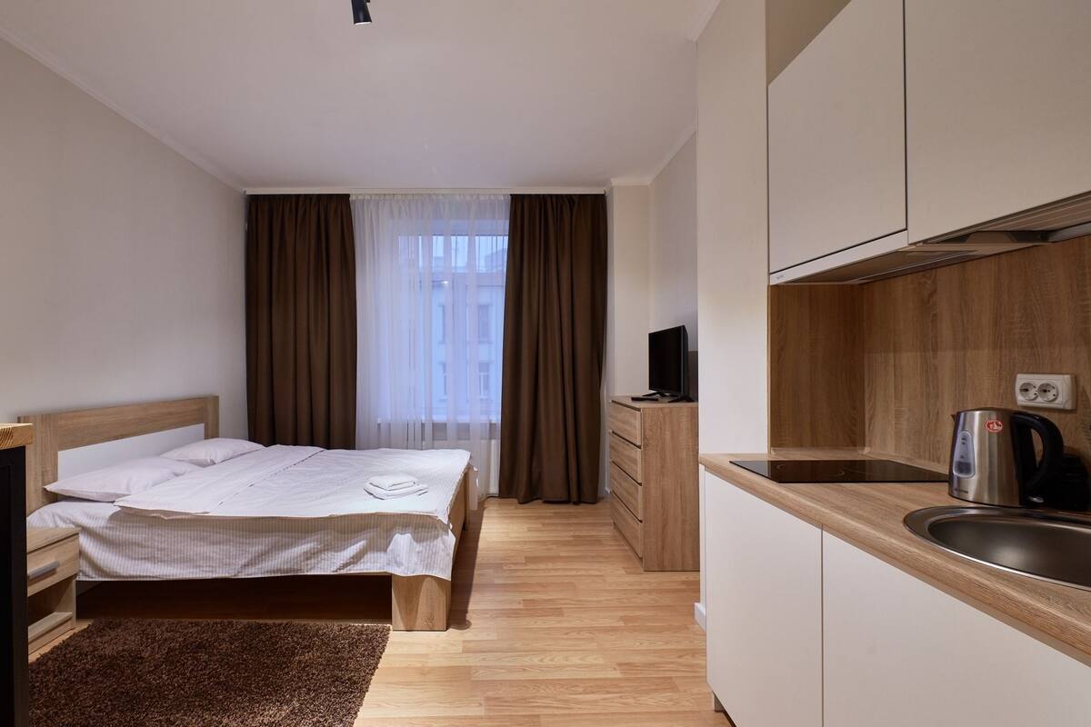 A hotel room with a bed, a small desk, a TV, a wooden cabinet, and a kitchenette with a kettle and a coffee maker.