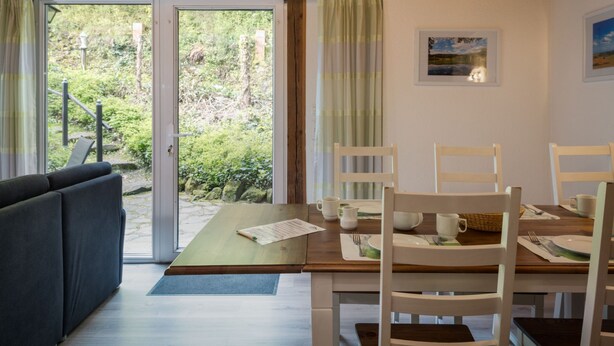 A dining area with a wooden table, white chairs, and a view of a forest through glass doors. - Eifelnatur (Haus 2) by Interhome (Immerath)