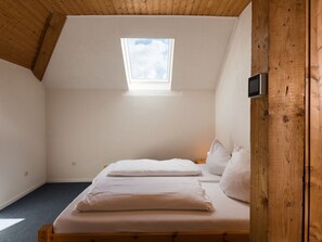 Quarto pequeno e acolhedor com teto inclinado e claraboia. Há uma cama com roupa de cama e almofadas brancas, e uma pequena cabeceira de madeira.