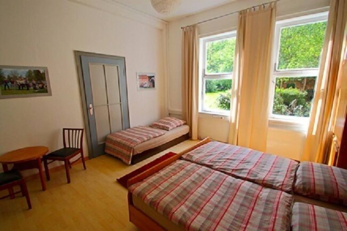 Un dormitorio con dos camas, una mesita pequeña y una silla. Hay una ventana con cortinas, una puerta y un cuadro en la pared.