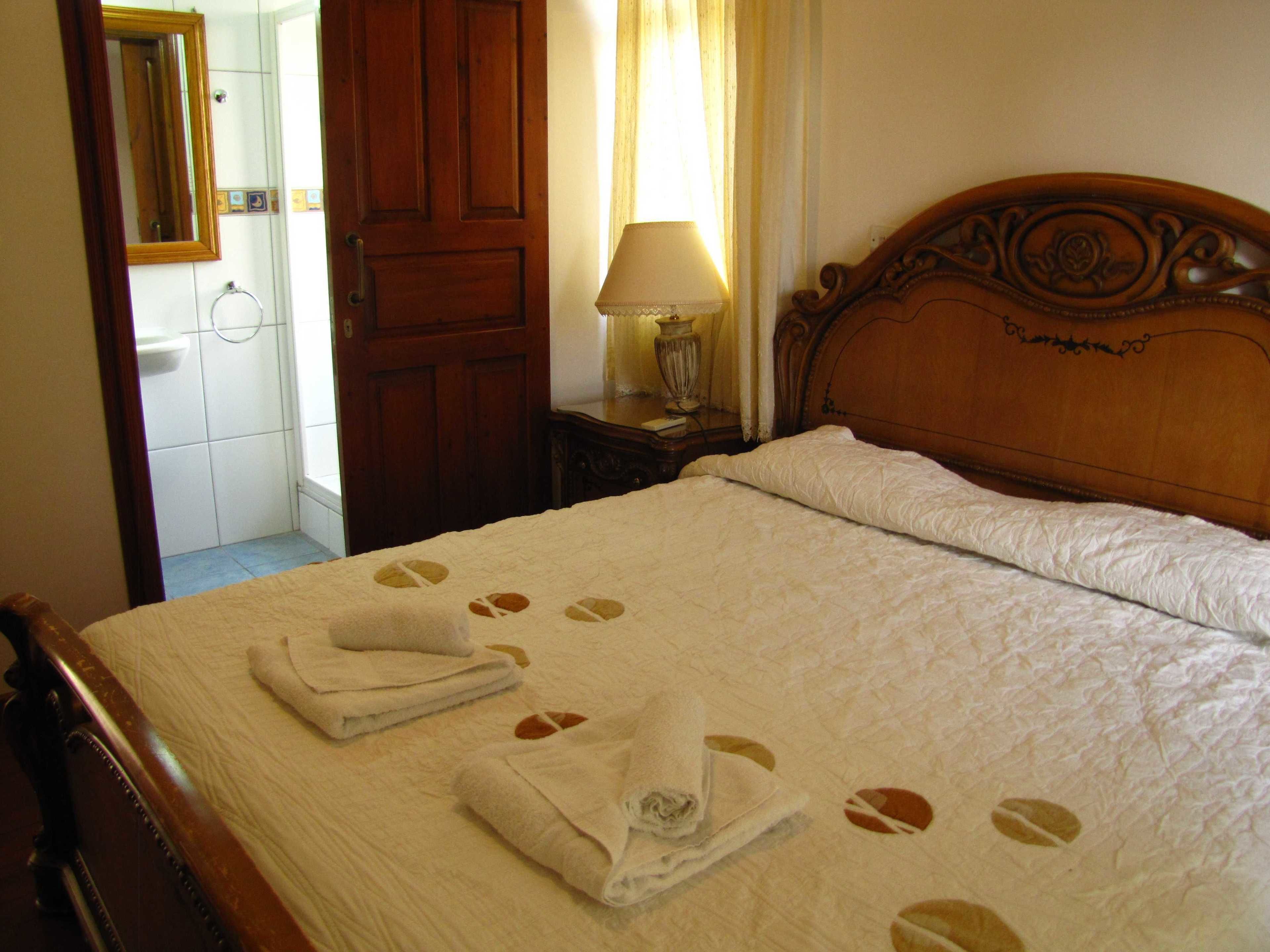 Una habitación de hotel con una cama de madera, una mesita de noche con lámpara y un baño visible a través de una puerta abierta.