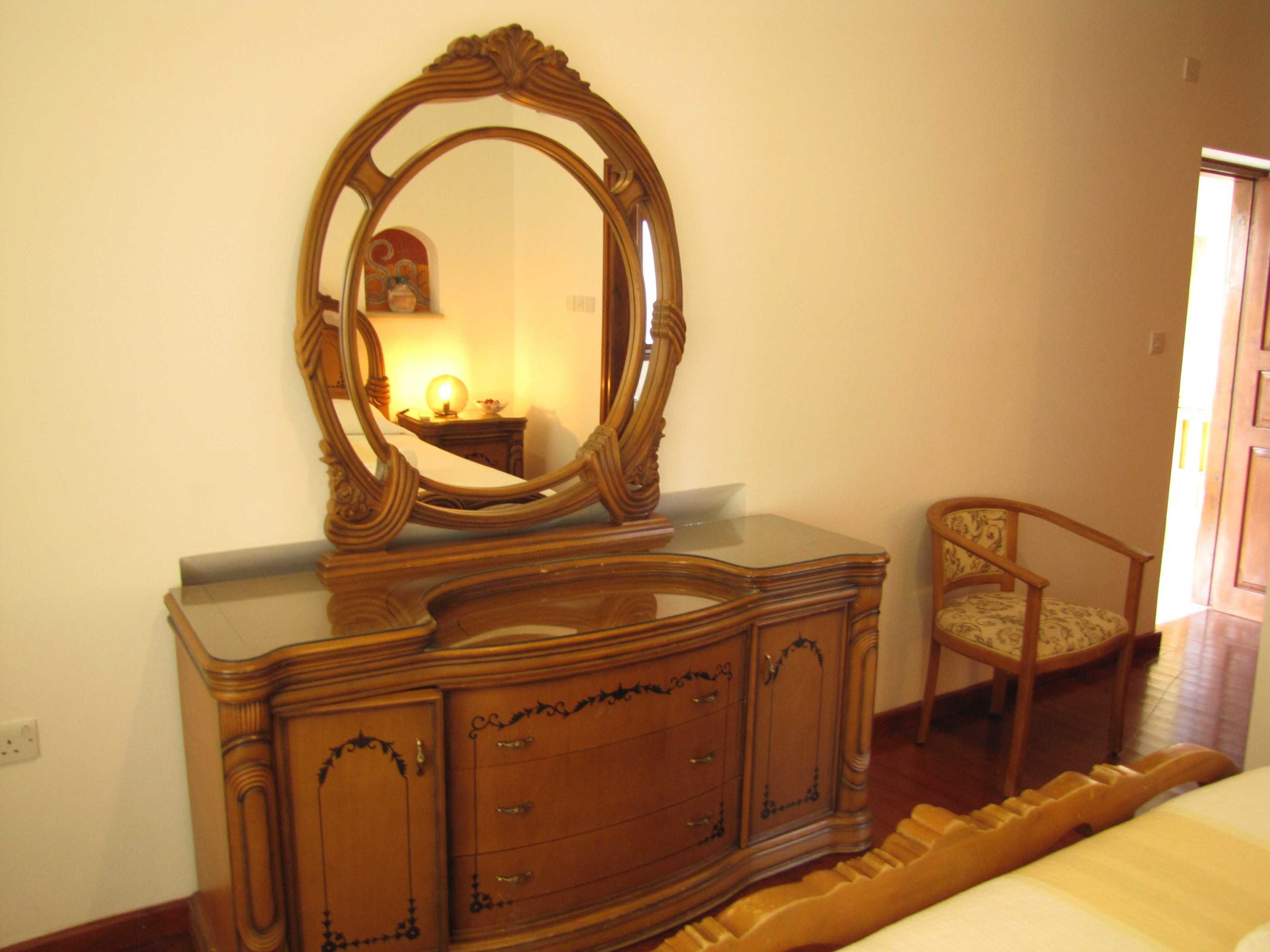 A wooden dresser with a large mirror, a chair, and a bedside table with a lamp.