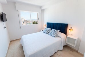 A bedroom with a bed, white walls, a window, a TV mounted on the wall, and a nightstand with a lamp.