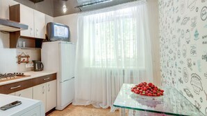 Eine kleine Küche mit einem Kühlschrank, einem Herd und einem Fernseher darüber. Ein Glastisch mit einer Schale Erdbeeren ist ebenfalls vorhanden.