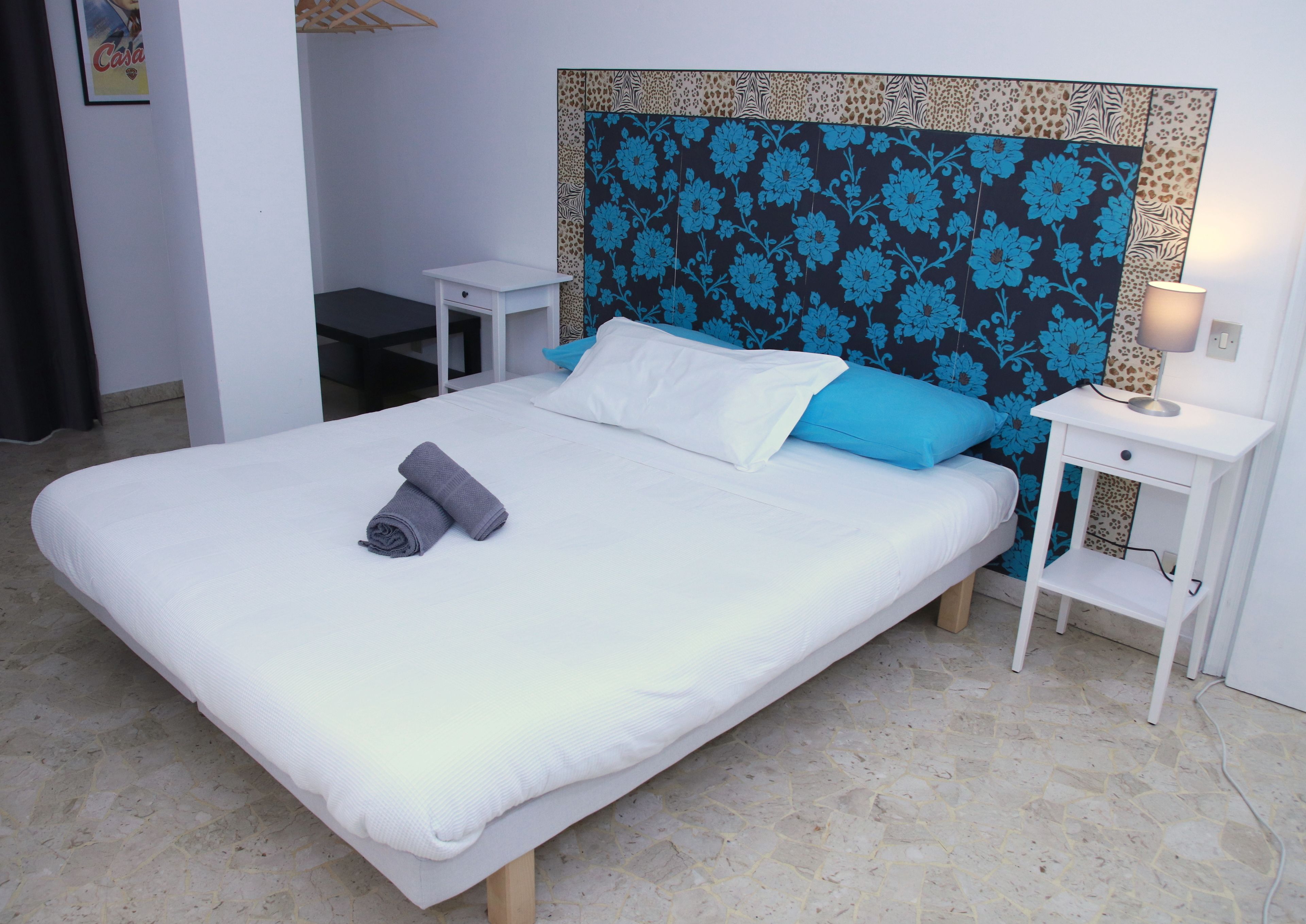 A neatly made bed with a white mattress, a blue floral headboard, and a grey towel. There are two bedside tables, one with a lamp and the other empty.