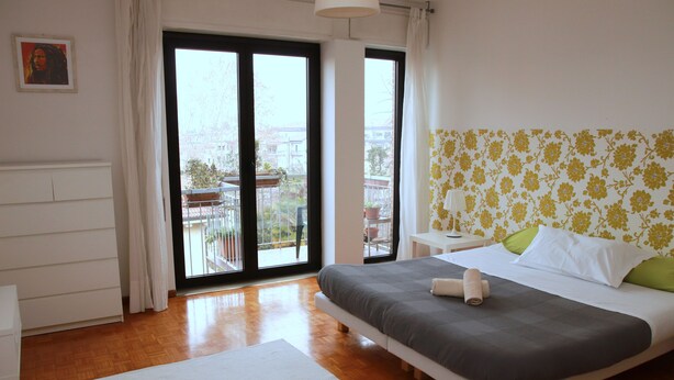 A bedroom with a large bed, a white nightstand, a white desk, and a view of the outside balcony. - Ca' Lou al Teatro (Vicenza)