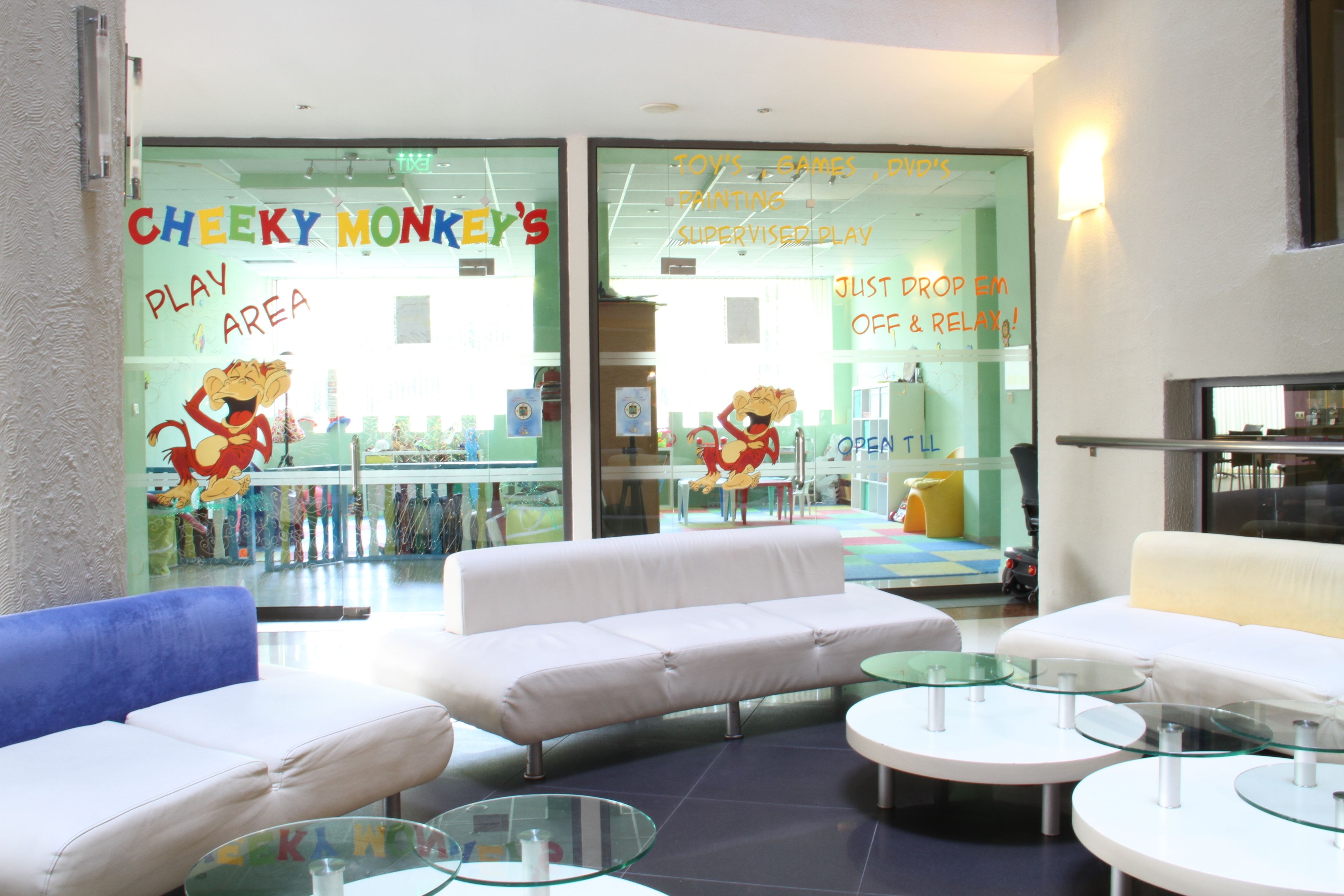 Espaço de recreação com marca Cheeky Monkey, sofás brancos e mesas de vidro.