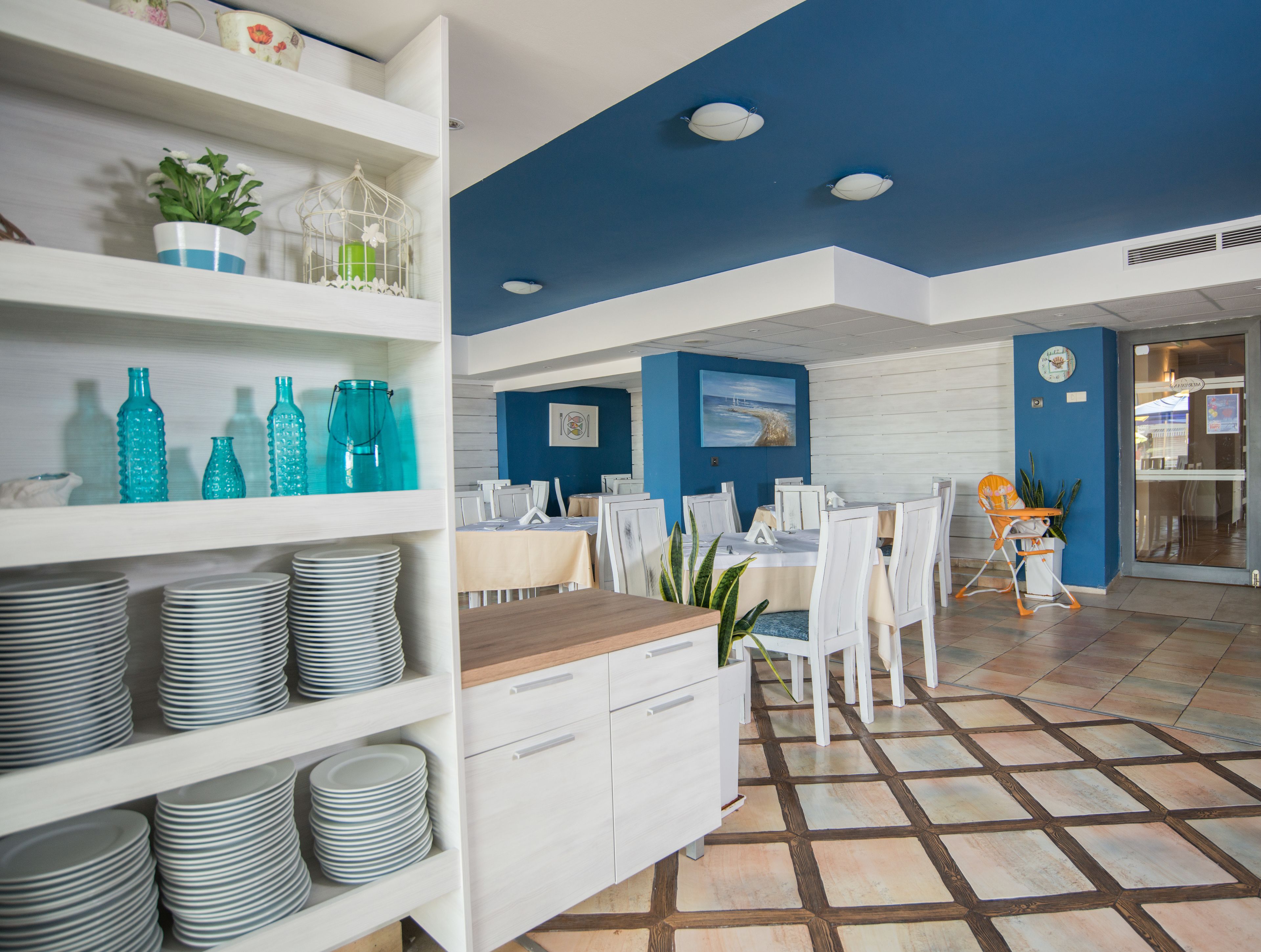 A modern dining area with white chairs and tables, a blue accent wall, and a tiled floor.