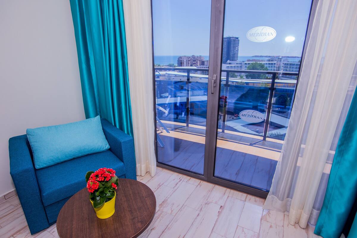 Una habitación moderna con un sofá azul, un jarrón amarillo con flores rojas y un ventanal con vistas a un paisaje urbano y una masa de agua.