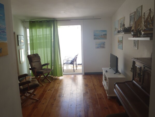 Uma sala de estar com piso de madeira, uma cortina verde, uma cadeira de balanço, um aparador de TV e um piano.