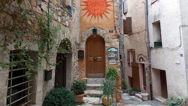 A narrow alley with a stone building featuring a large sun mural, a wooden door, and potted plants. - Nice little studio 20 M2 (AUPS)