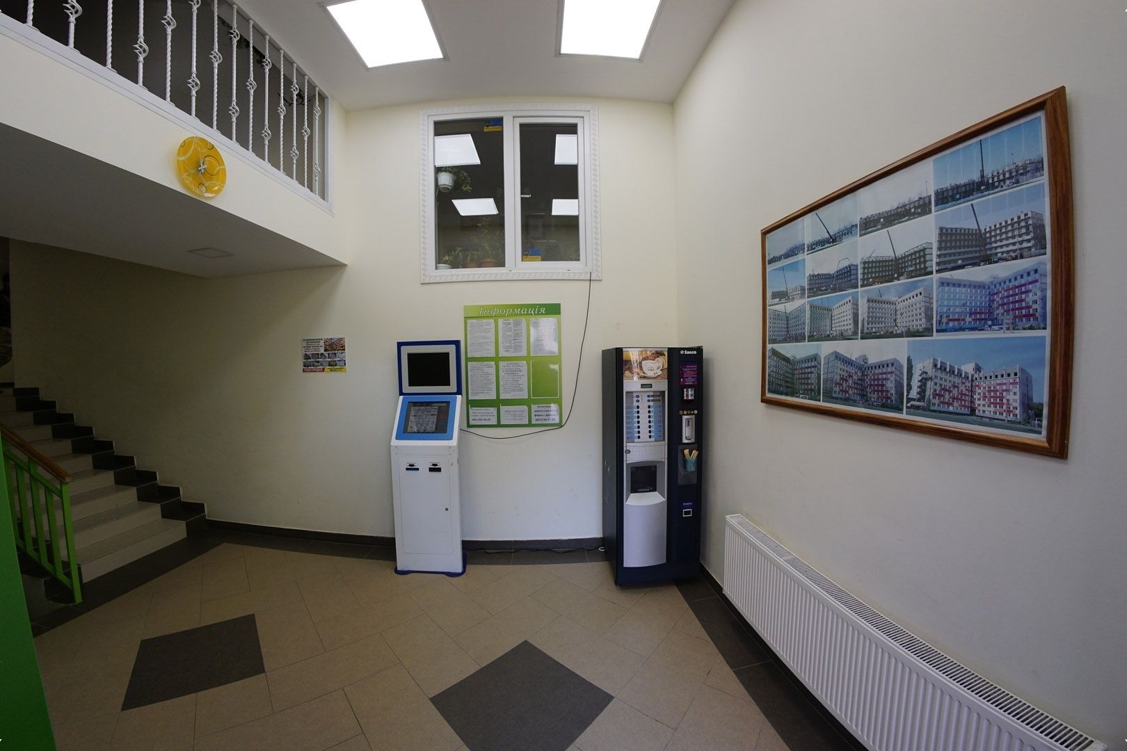 A lobby with a staircase, a self-service kiosk, a radiator, and a framed cityscape photo.