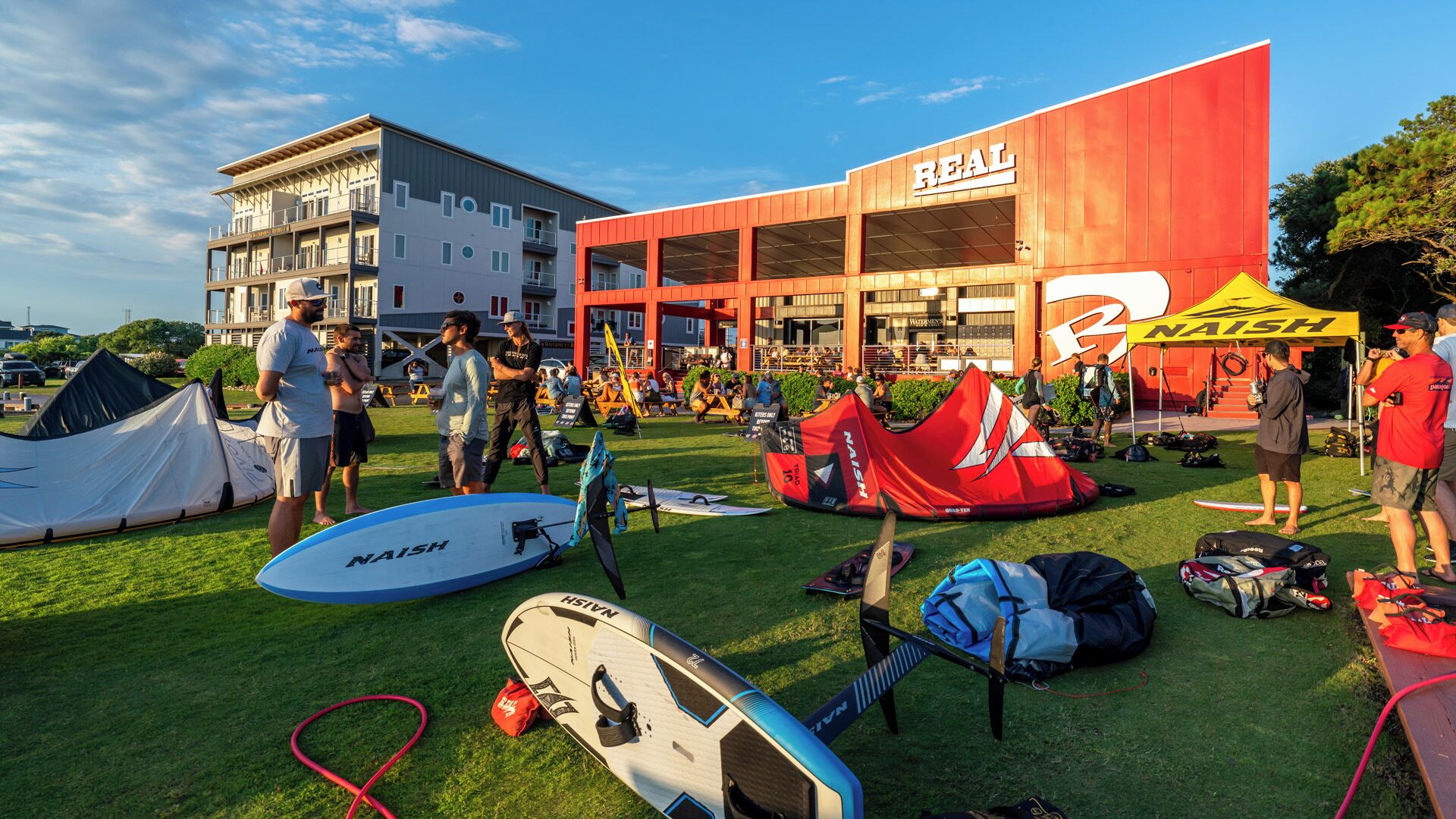 A building with a 'REAL' sign, a grassy area with surfboards, and tents.