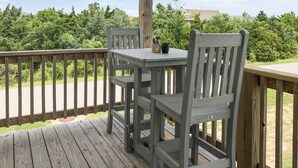 A wooden deck with a table and chairs, surrounded by greenery and a clear sky. - 3-bedroom, 2-bathroom apartment. Walk to beach and access to REAL Watersports (Waves)
