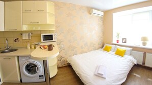 Een compacte hotelkamer met een kitchenette, een wasmachine, een bed met witte en gele kussens, en een raam met uitzicht.