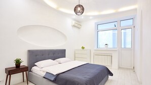 Een moderne slaapkamer met een grijs gestoffeerd bed, een nachtkastje met een plant, een radiator en een raam met jaloezieën.