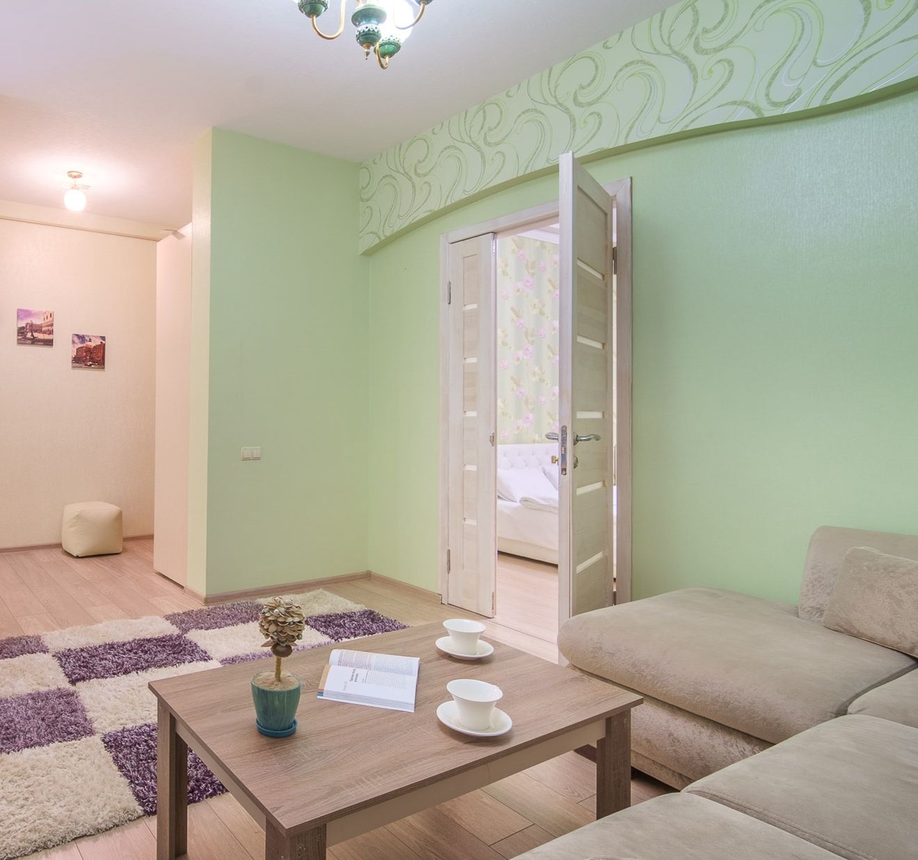 Una sala de estar con un sofá, una mesa de centro con un florero y tazas, una puerta que da a un dormitorio, y una alfombra estampada.