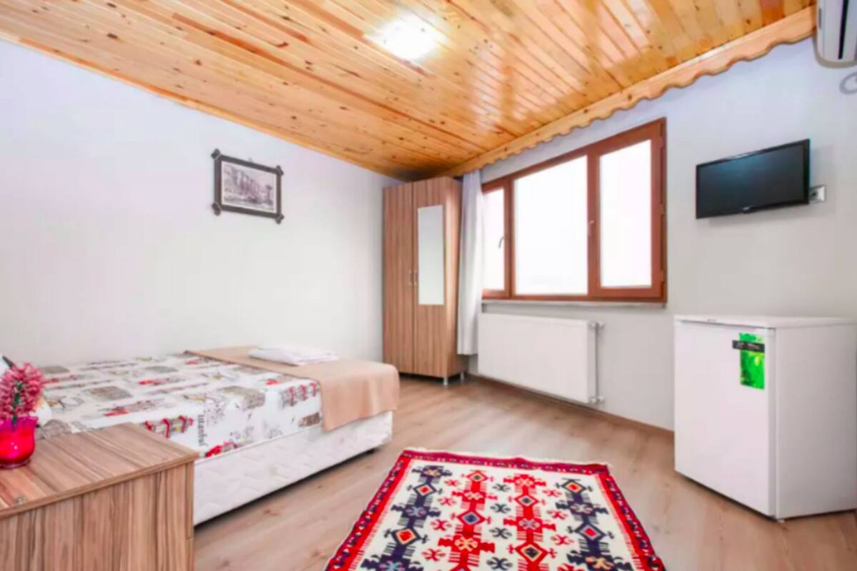 A bedroom with a bed, a small table with a vase, a television, a window, and a refrigerator.