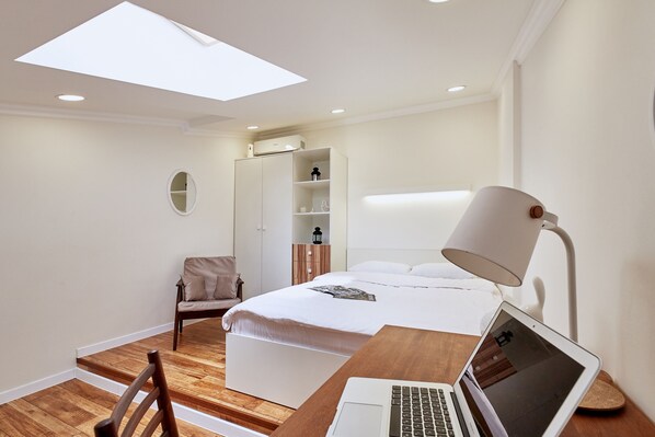 A modern bedroom with a bed, a desk with a laptop, a chair, a bedside lamp, and a skylight.