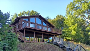 Eine zweigeschossige Holzhütte mit einem Balkon, umgeben von Bäumen und einem grasbewachsenen Hügel.