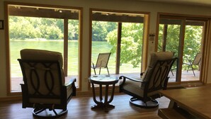 Ein Zimmer mit zwei Stühlen und einem Tisch, große Glasüren führen zu einer Terrasse mit Blick auf einen See und Bäume.