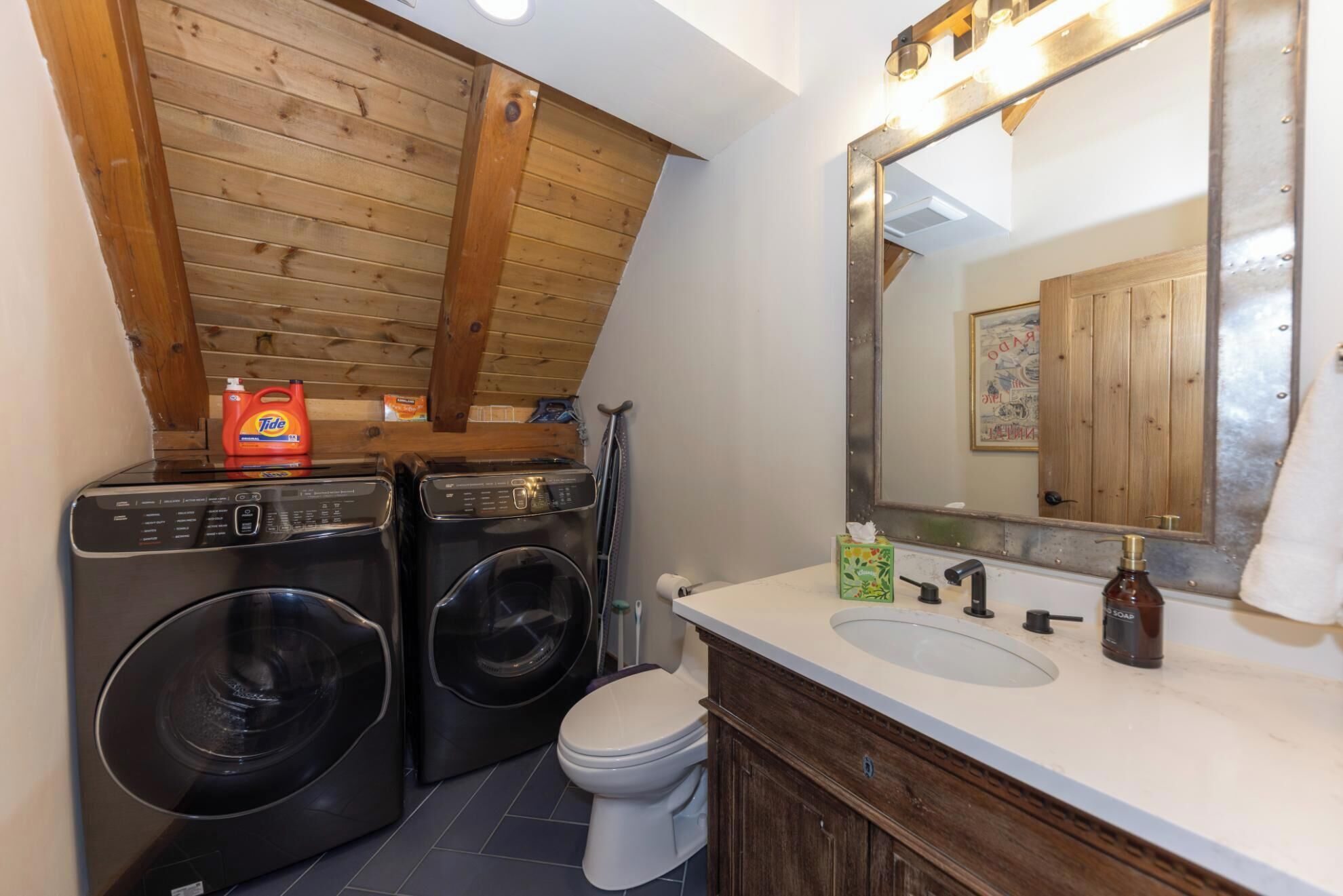 A compact bathroom with a wooden ceiling, two front-loading washing machines, a toilet, a sink with a mirror, and a small shelf with toiletries.