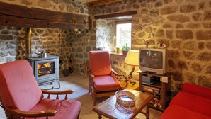 Une pièce rustique aux murs de pierre, avec un plafond en bois, une cheminée, deux fauteuils rouges, un téléviseur et une petite table sur laquelle se trouve un panier.