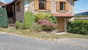 Una casa de piedra con contraventanas de madera, un pequeño porche y un jardín lleno de flores rosadas.