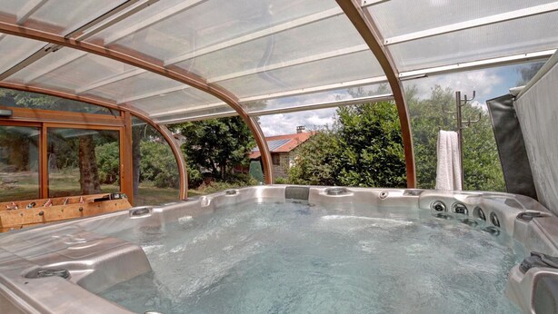Tinaja de hidromasaje con recinto de vidrio, rodeada por una terraza de madera y vistas a una casa y árboles.