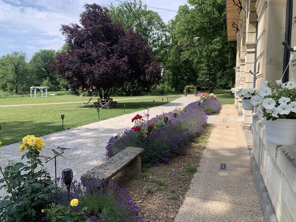 A garden pathway with benches and flower beds, a pergola, and a building with hanging flower pots.
