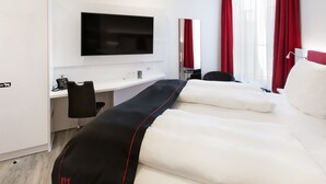 Una habitación de hotel moderna con televisión de pantalla plana en la pared, un escritorio con teléfono, y una cama con ropa blanca y detalles en rojo y negro.