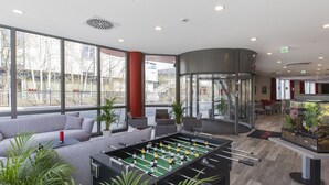 Un vestíbulo moderno con mesa de fútbol, asientos cómodos, ventanas amplias y un ascensor de vidrio.