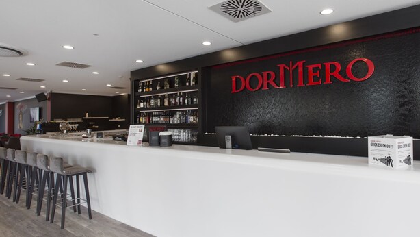 Un área de bar moderna con una pared negra y letras rojas que dicen 'DORMERO'. Hay taburetes de bar, una computadora portátil y un cartel que dice 'A TANCY WELCOME'.