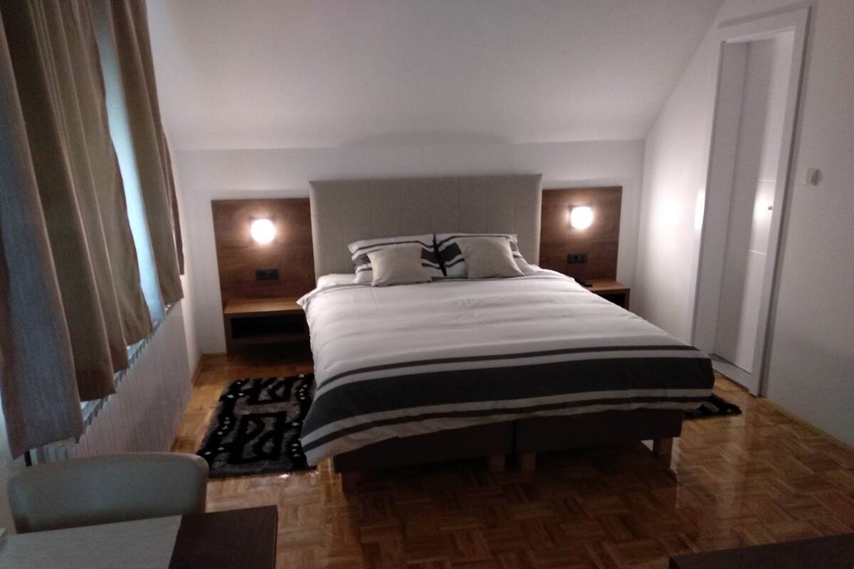 A bedroom with a large bed, two bedside tables, a nightstand, a chair, and a wooden floor.